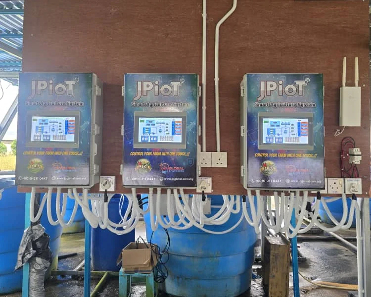 Three JPIoT Smart Agriculture System controllers with touchscreens, mounted side by side on a wooden board, with white conduits running down to blue water tanks