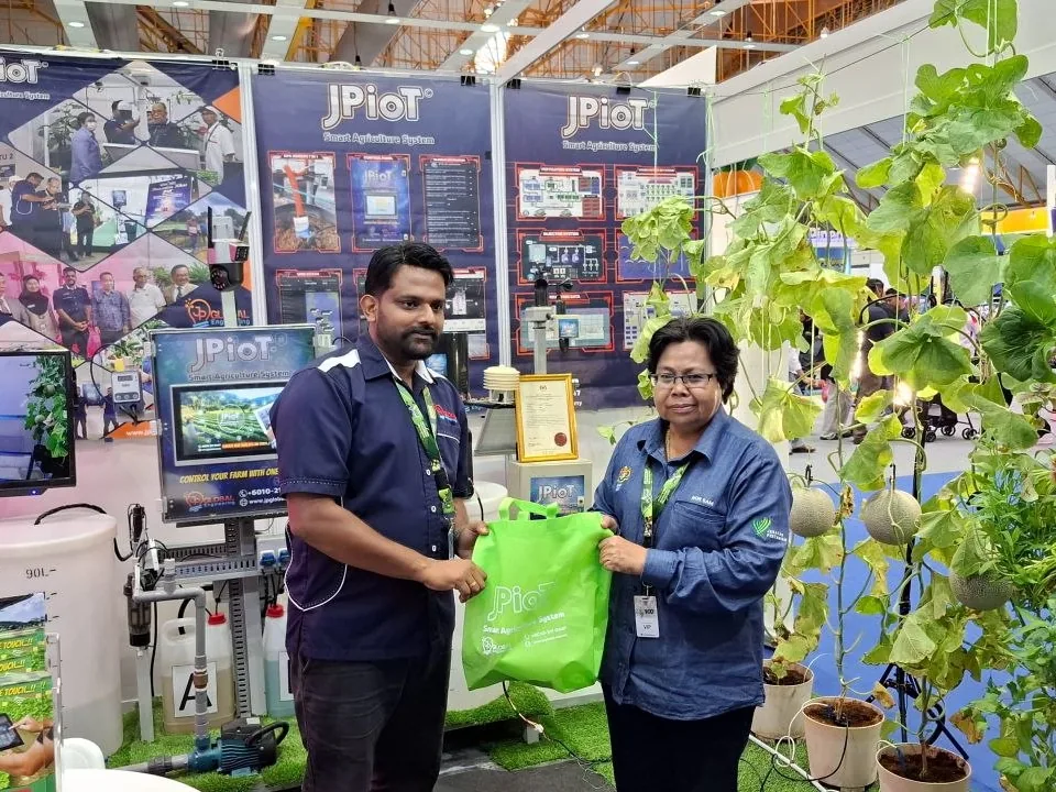 At the JPIoT stand, a JP Global team member hands a green JPIoT bag to a guest wearing a VIP pass