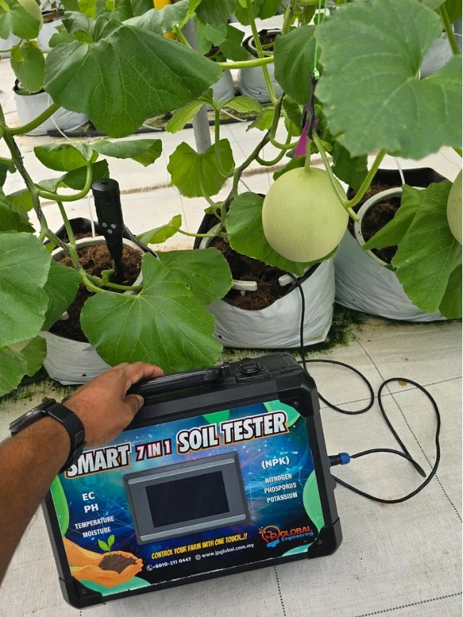 The tester on a woven ground sheet beside melon plants growing in grow bags, a hand resting on the case and the cable running from its side